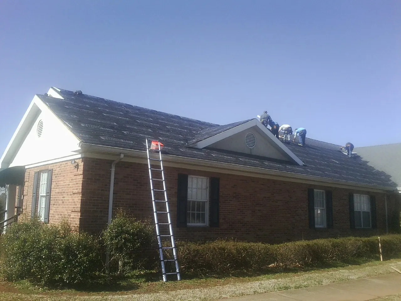 Crew tearing shingles off a brick ranch roof with a ladder set — Emergency Roof Repair work by our crew in Gillsville