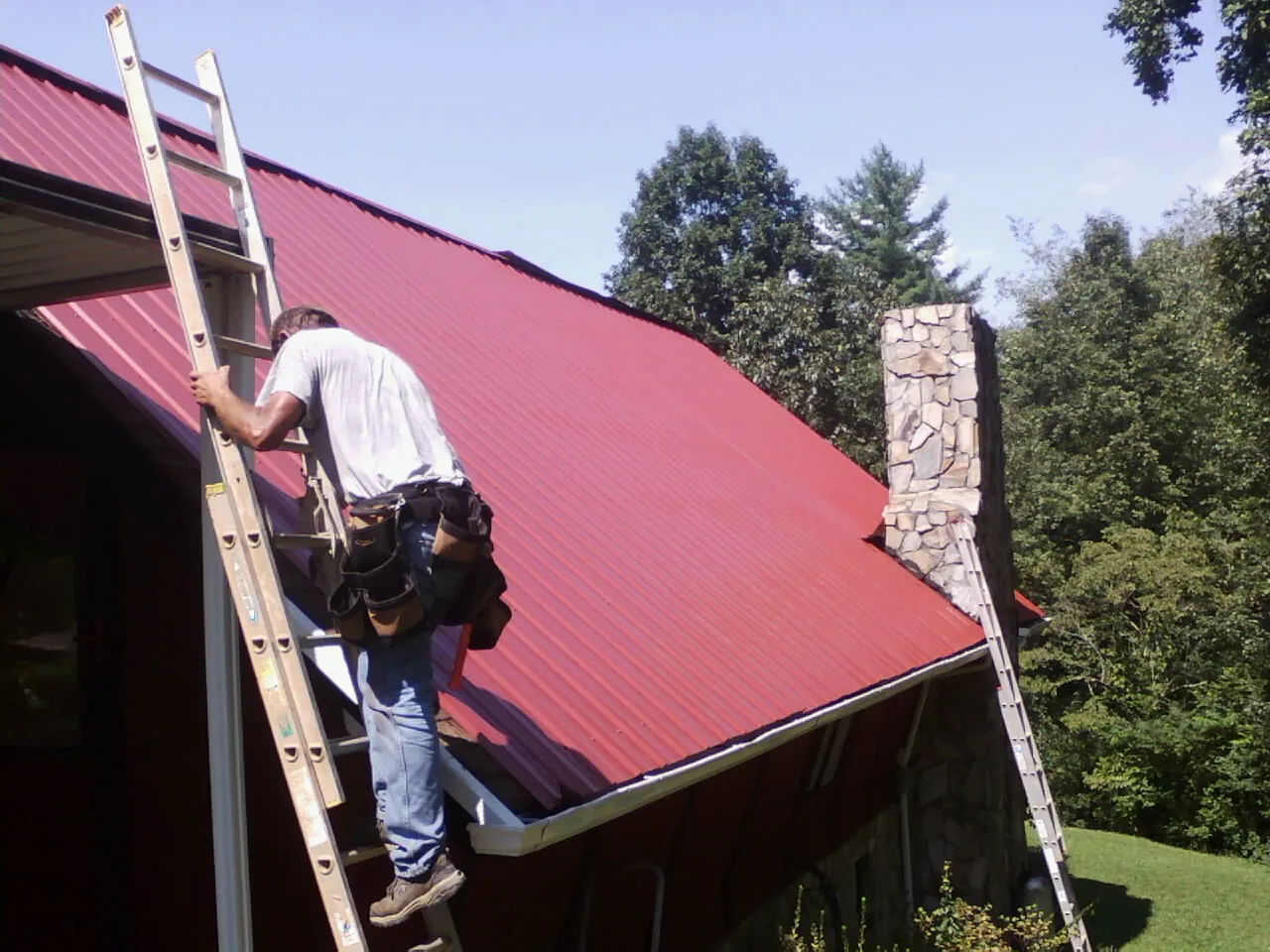 Roofer on a ladder beside a red metal roof and stone chimney — Soffit & Fascia Repair by Roofing Brothers in Gillsville